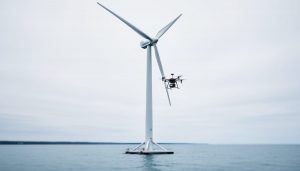 Modern wind turbine near the Ontario shoreline with a nearby inspection drone and a floating offshore platform structure in the background.